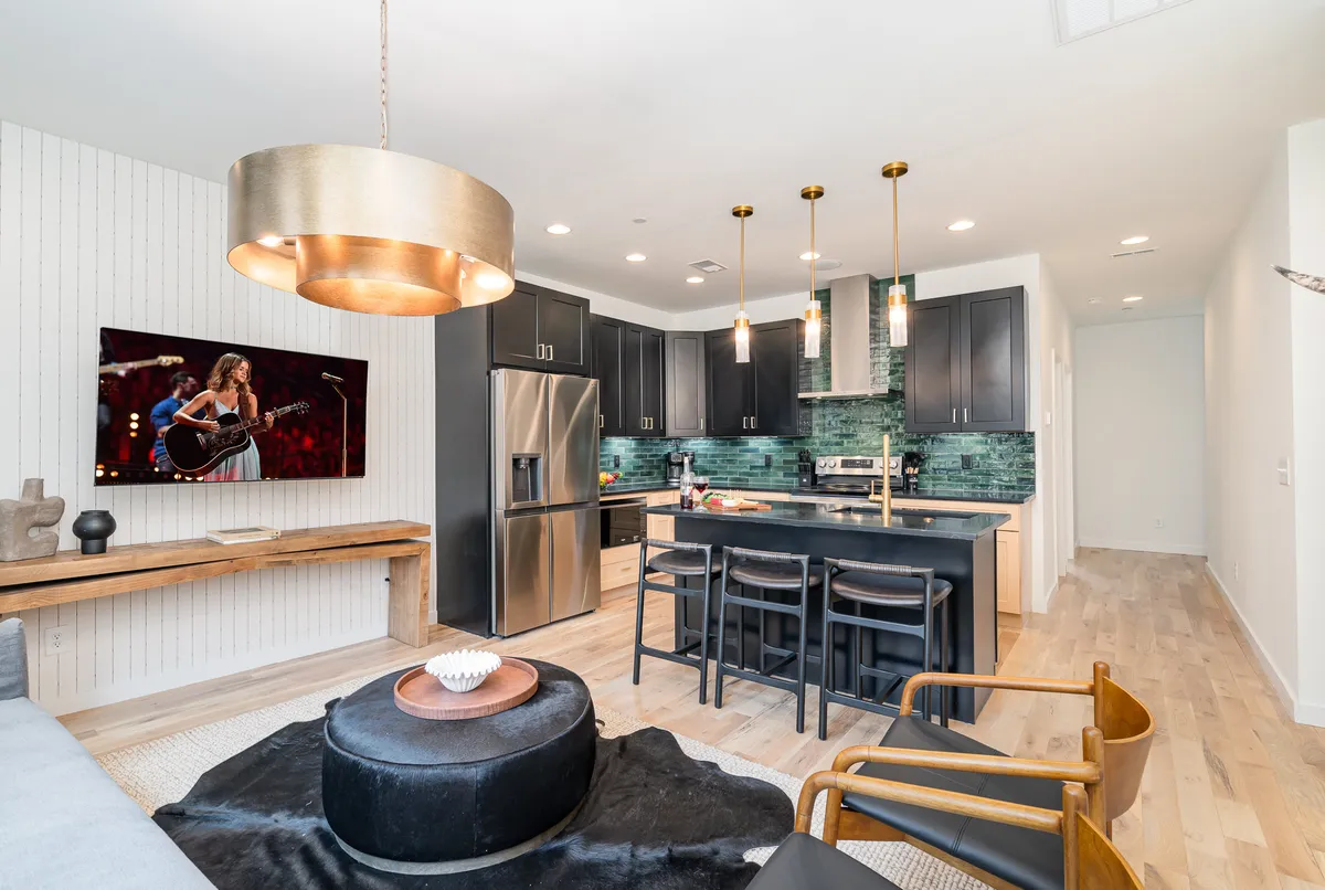 The image showcases a spacious, modern kitchen with a large island perfect for gathering. Sleek black cabinetry and elegant pendant lighting set a stylish tone, while a TV displaying a live artist performance hints at Nashville's music scene. The layout invites conversation and connection, ideal for cooking, entertaining, or relaxing with coffee in the morning.