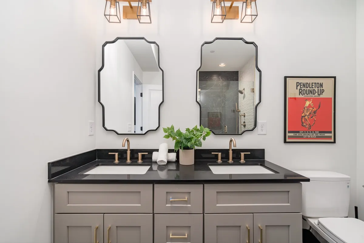 A modern bathroom showcasing double vanities with stylish sinks, sleek mirrors, pendant lights, a touch of greenery, and a framed poster. The space is bright, fresh, and designed for comfort and convenience.