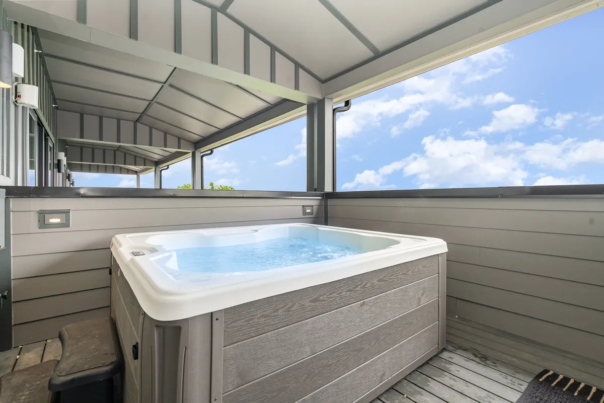 A beautiful rooftop deck featuring a bubbling hot tub, warm wooden decking, and lush surrounding greenery. The space is open to the sky, inviting relaxation and socializing with friends while enjoying panoramic views, possibly at sunset or under the stars.