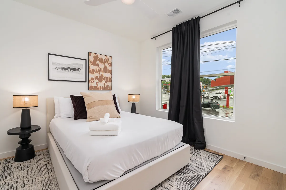 A serene bedroom featuring a bed with crisp white linens, stylish artwork above the headboard, nightstands with matching lamps, and a large window that allows natural light to flood the space. The overall atmosphere is calm, inviting, and ideal for relaxation.