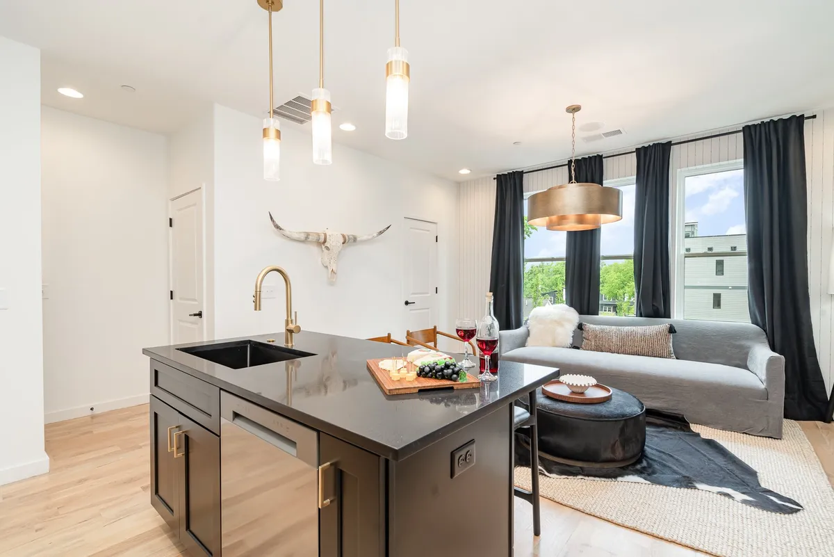 A cozy and stylish open-plan kitchen and living area centered on a dark kitchen island topped with a cheese platter and wine, surrounded by charming décor. Visible are a comfortable sofa, decorative pillows, and large windows, evoking a warm, communal atmosphere ideal for gatherings.
