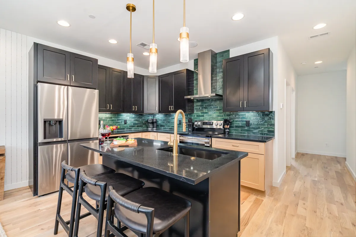 A modern kitchen interior featuring a central island, gleaming stainless steel appliances, a striking green backsplash, and elegant gold pendant lights overhead. The kitchen island offers bar seating, and the space is styled to feel both welcoming and practical—ideal for cooking and socializing.