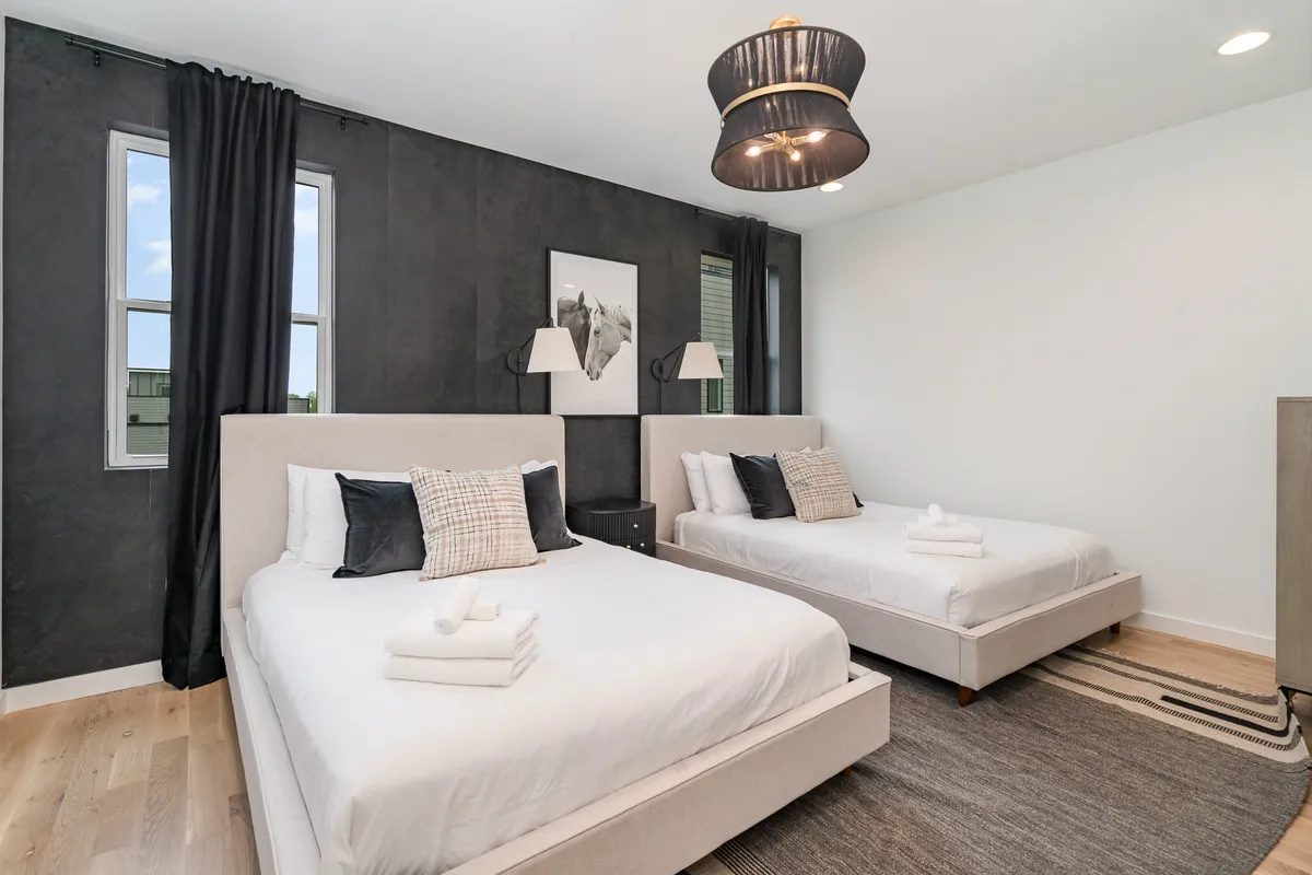 A spacious bedroom with two beds dressed in soft, neutral linens, a dramatic black accent wall, a large pendant light fixture, and tasteful framed artwork. The room is bathed in natural light, creating a warm and inviting space perfect for unwinding or sharing with family or friends.