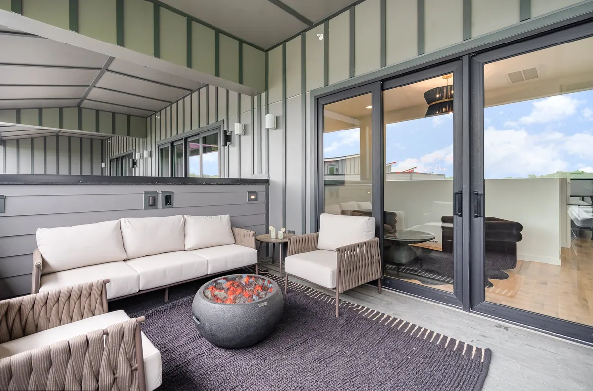 A cozy rooftop deck with comfortable seating gathered around a glowing fire pit. Large glass doors connect the space to the indoors, while the setting sun hints at city views and a welcoming space to unwind after a night out.