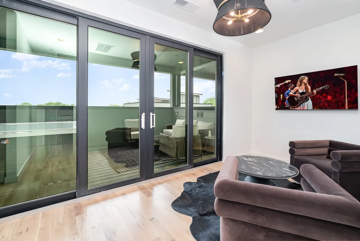 A contemporary living space opens to a balcony through large sliding doors, featuring comfortable seating, a TV showing a live performance, and plenty of natural light. The space invites both relaxation and collaboration, bridging work and leisure.